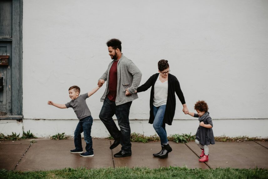 A man, woman and 2 kids all holding hands walking down a wet pavement.