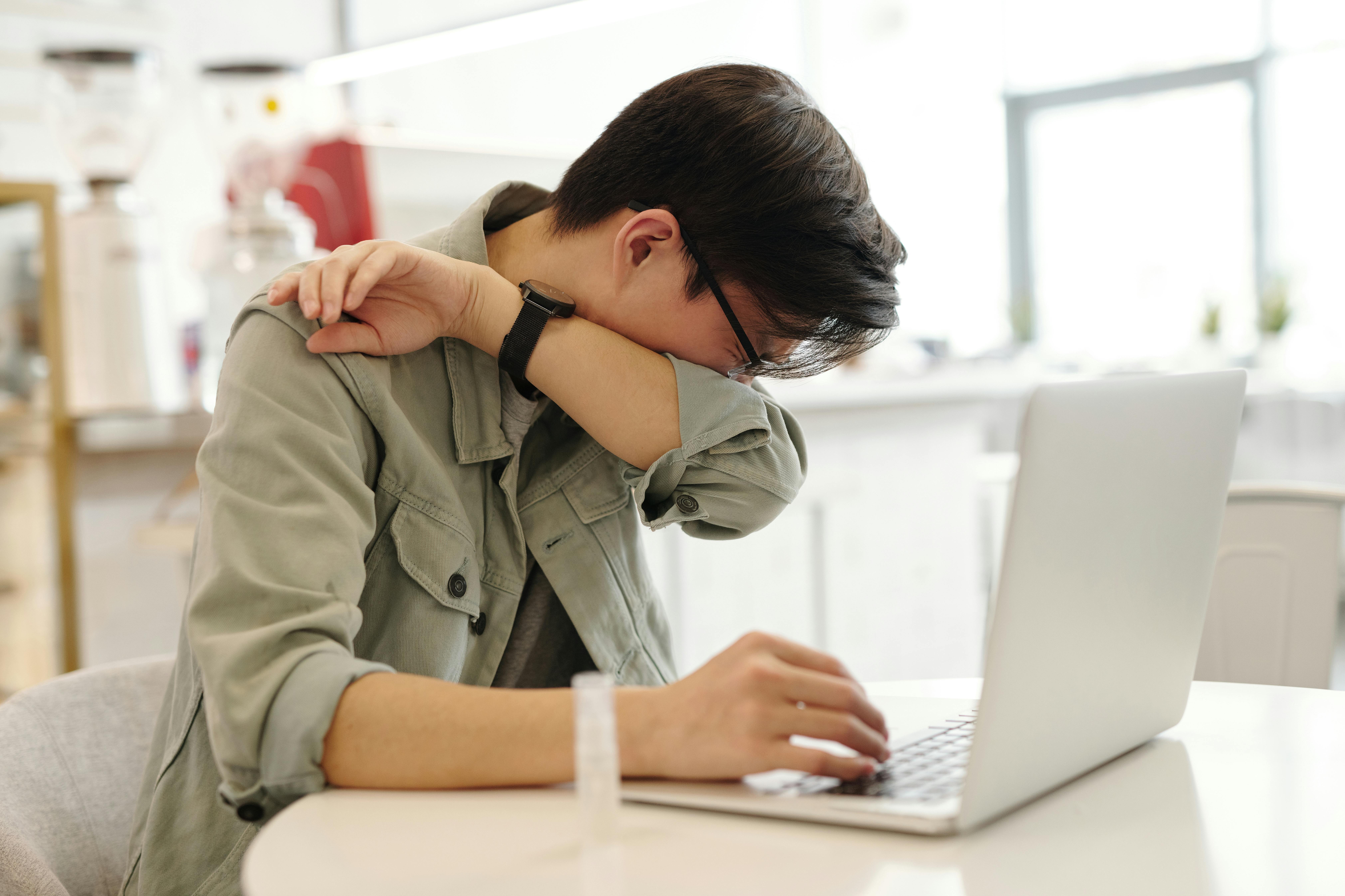A man sitting at a table with his laptop, sneezing into his elbow.